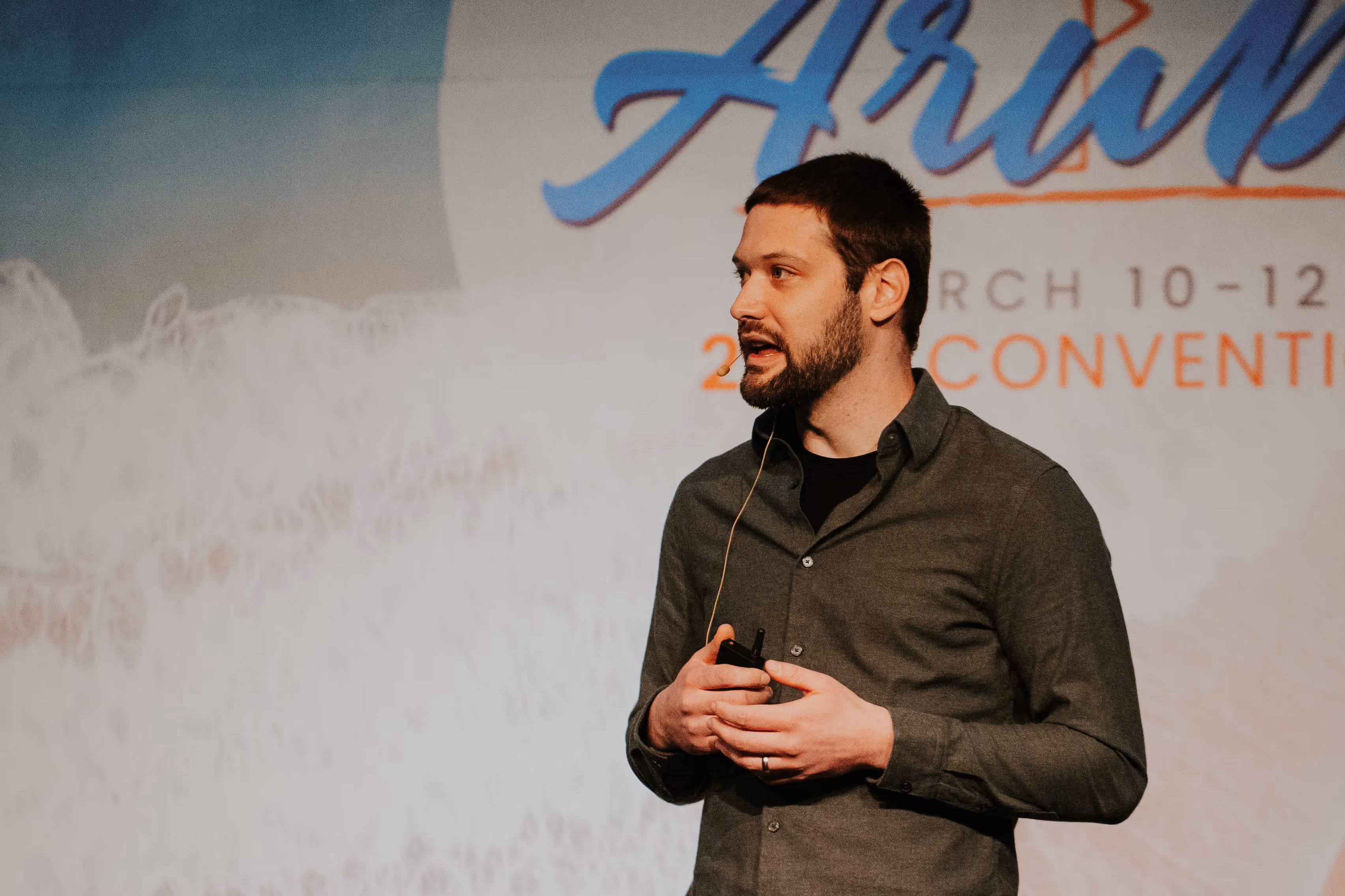 Zach with a beard wearing a headset microphone speaks on stage while holding a presentation clicker, with a large convention backdrop behind him.