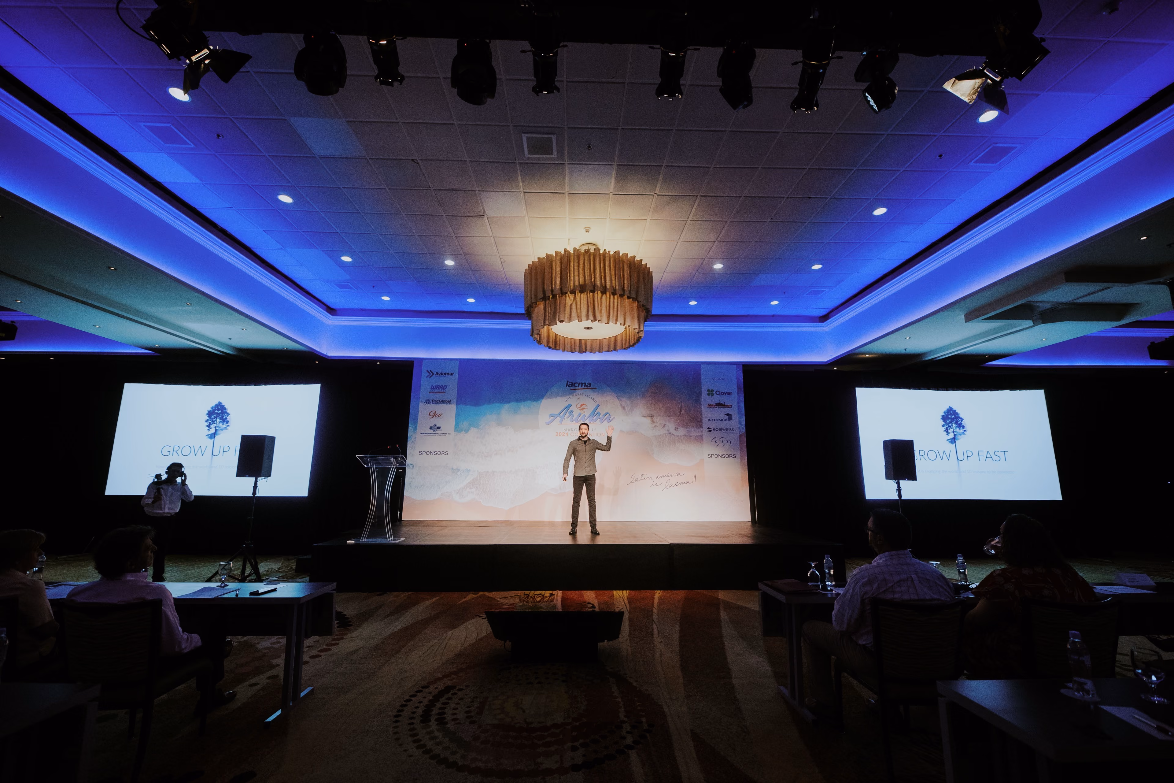 A wide shot of a speaker on stage at a corporate conference in Aruba, waving to an audience seated in a dimly lit ballroom with blue ambient lighting and presentation screens.