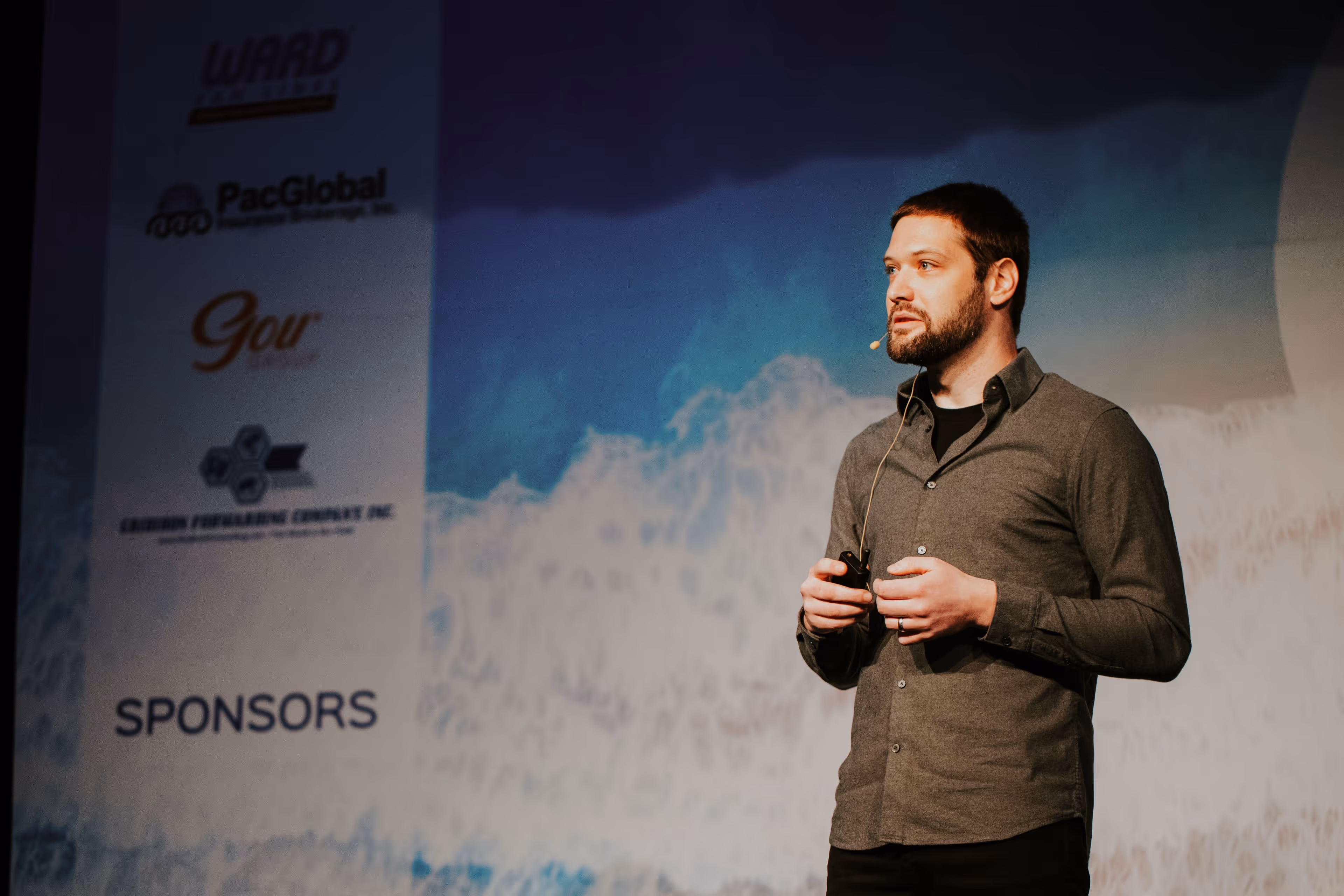 Zach giving a presentation on stage wearing a grey button-down shirt and a headset microphone, with a large presentation screen displaying sponsor logos in the background.