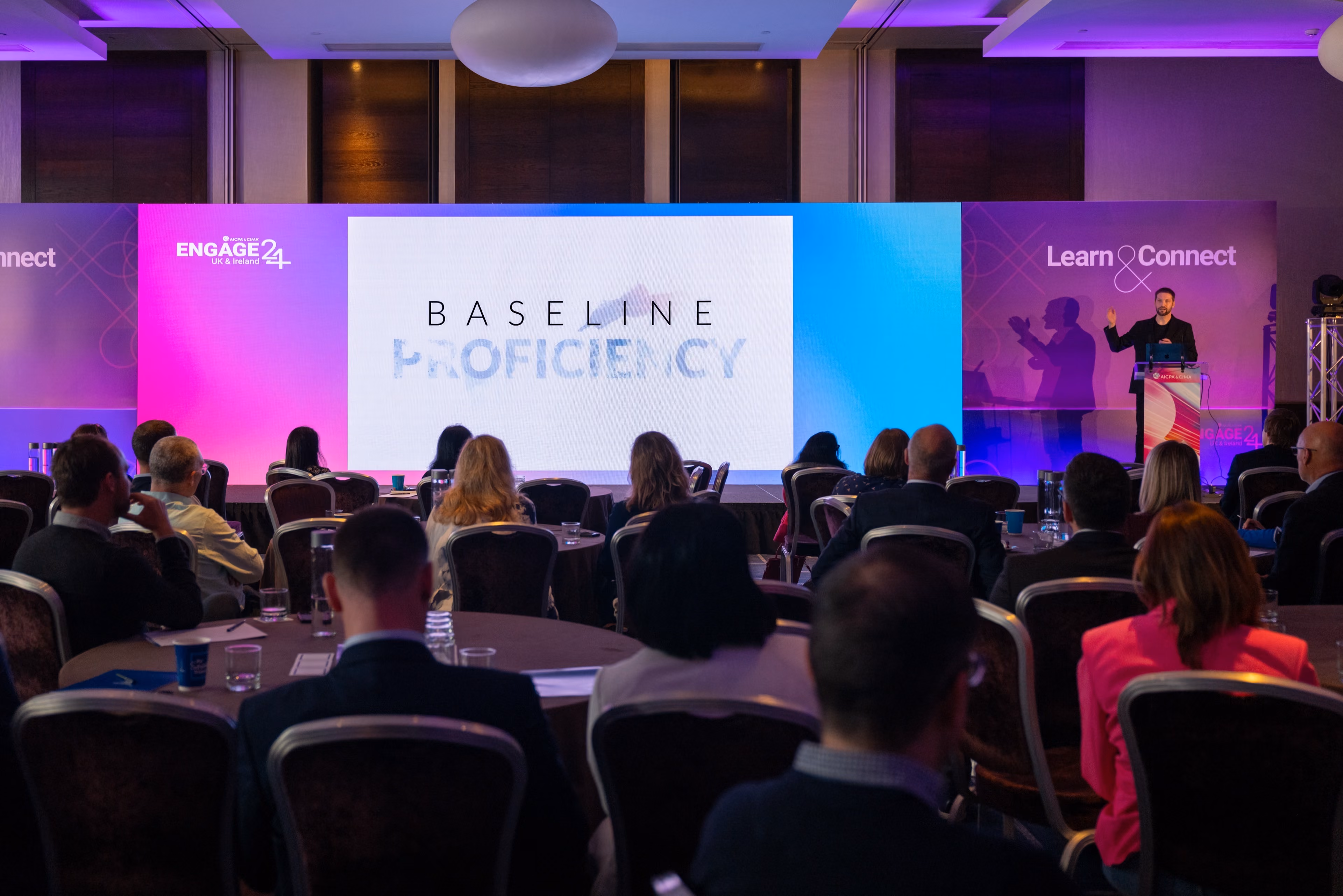 A speaker presenting on stage at a brightly lit corporate event to an audience seated at round tables, with a large screen displaying the words BASELINE PROFICIENCY.