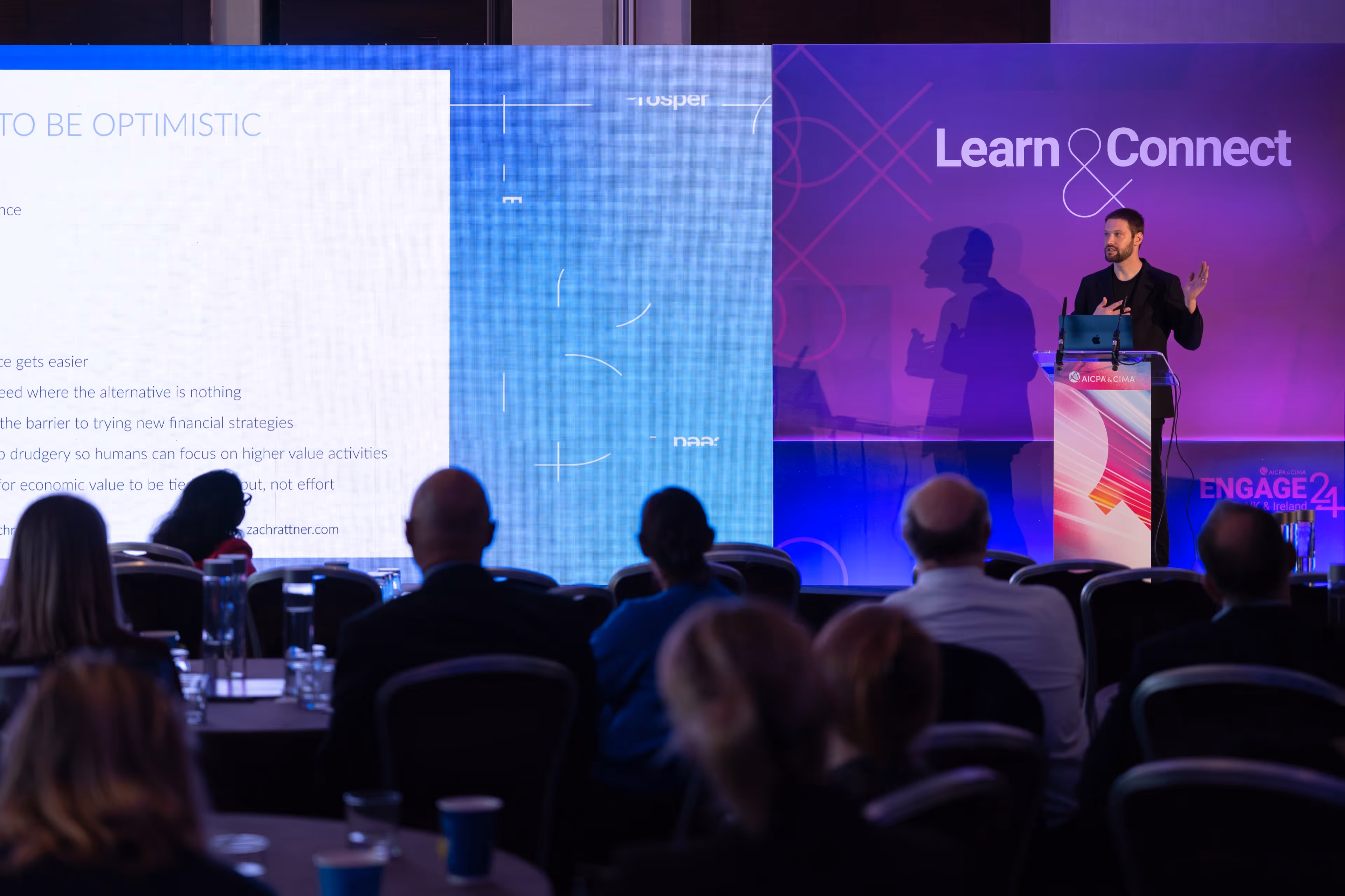 A male speaker presenting on stage at a corporate conference to a seated audience, with a large slide displaying the text TO BE OPTIMISTIC.