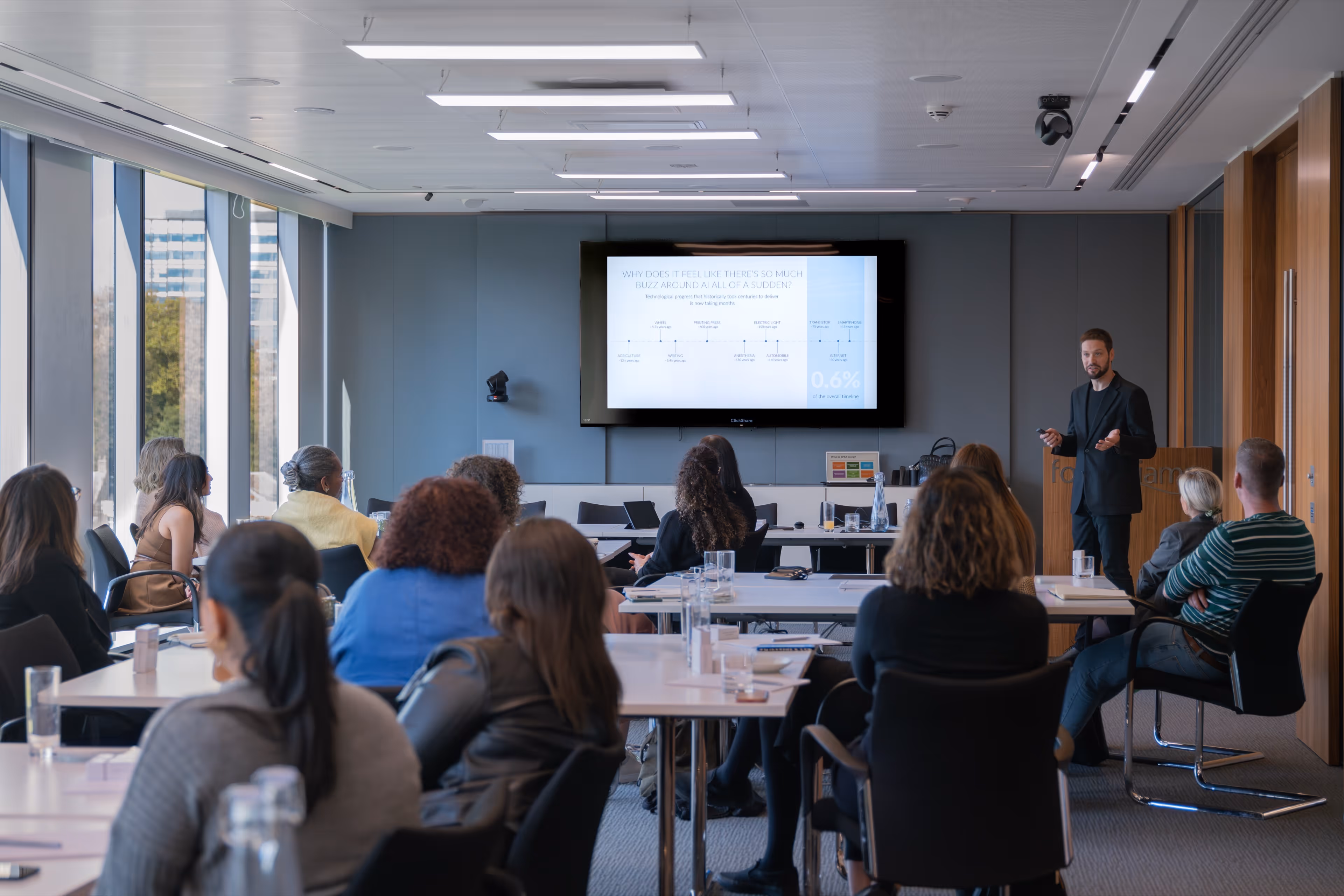 A male speaker giving a presentation about AI to an attentive audience seated around tables in a modern corporate boardroom.