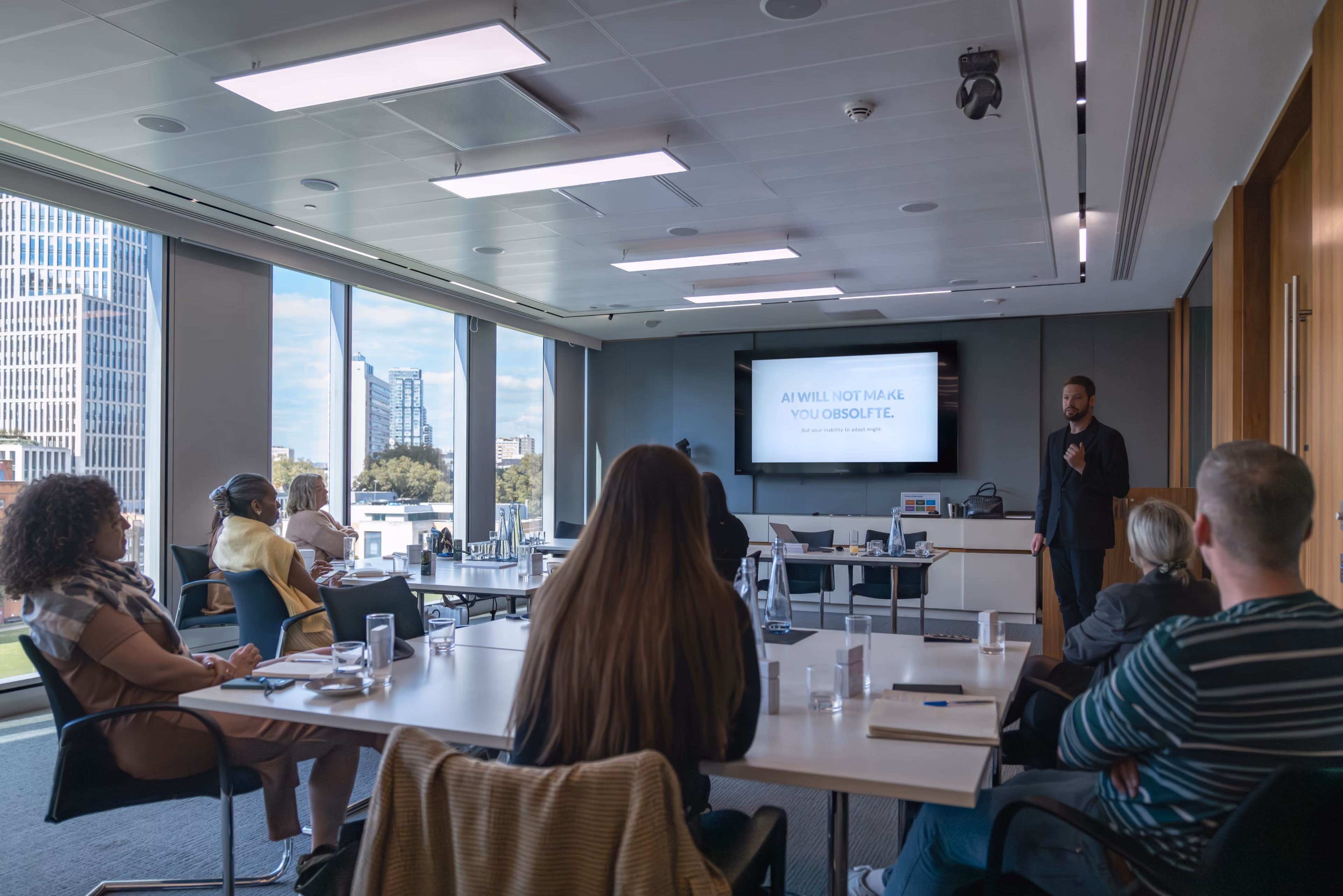 Zach giving a presentation about AI to an attentive seated audience in a modern corporate conference room with large windows overlooking a city.