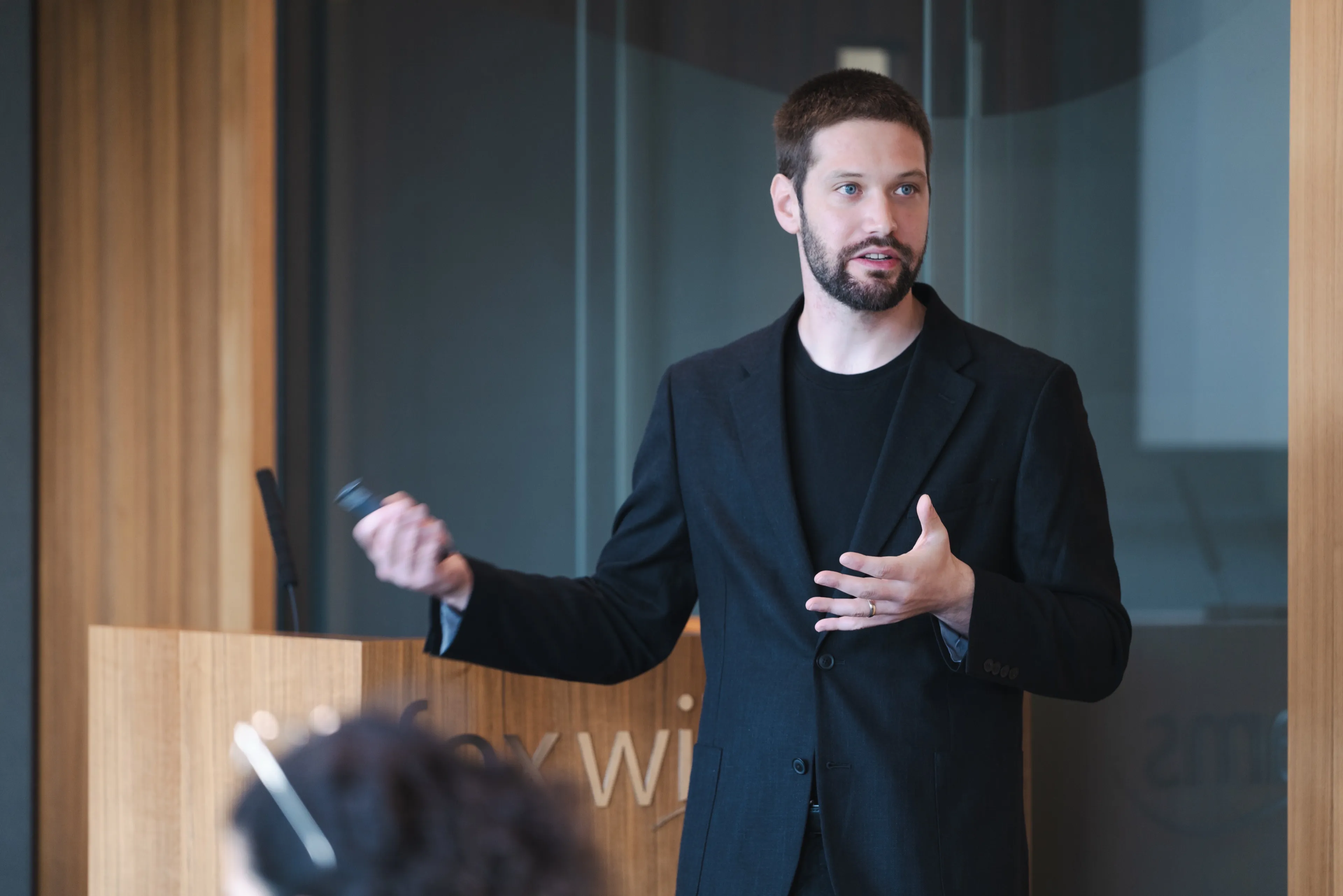 A male professional in a black blazer and t-shirt is presenting at a wooden podium, holding a presentation clicker and gesturing while addressing an audience.