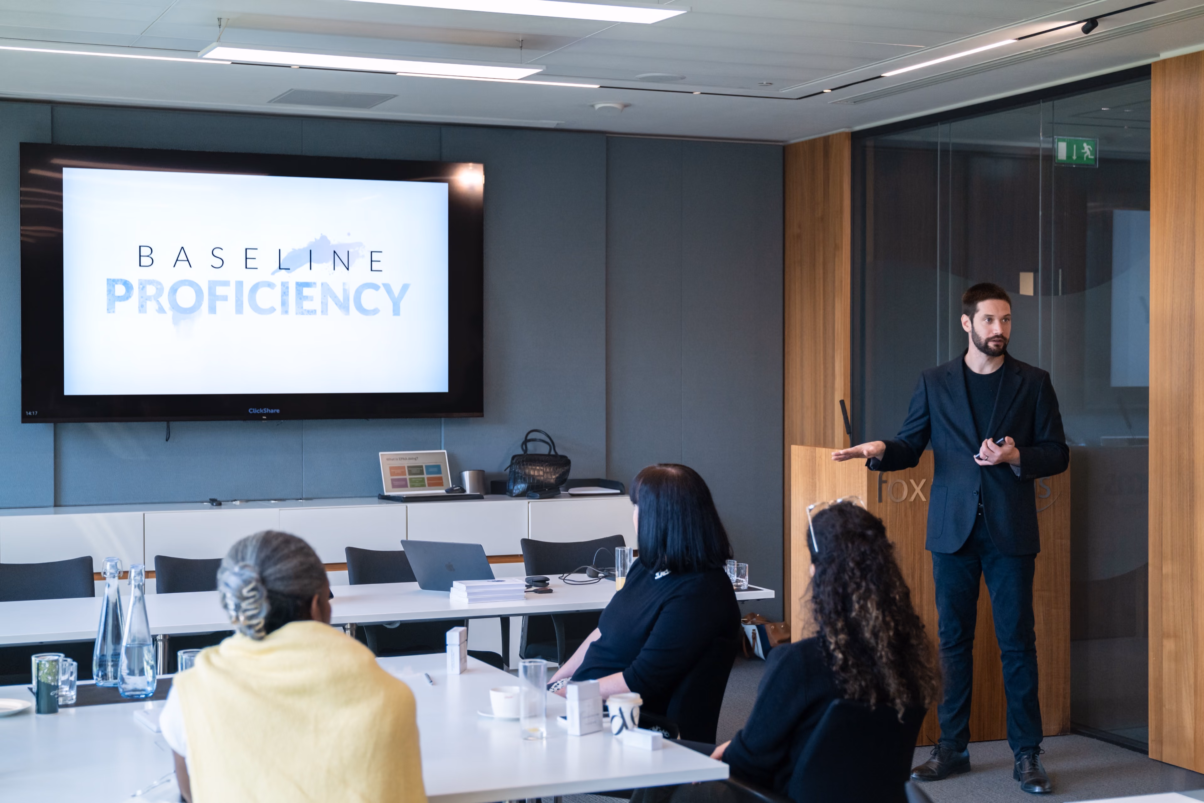 Zach professional in a dark blazer presenting to a small group of seated attendees in a modern conference room, with a large screen displaying BASELINE PROFICIENCY.