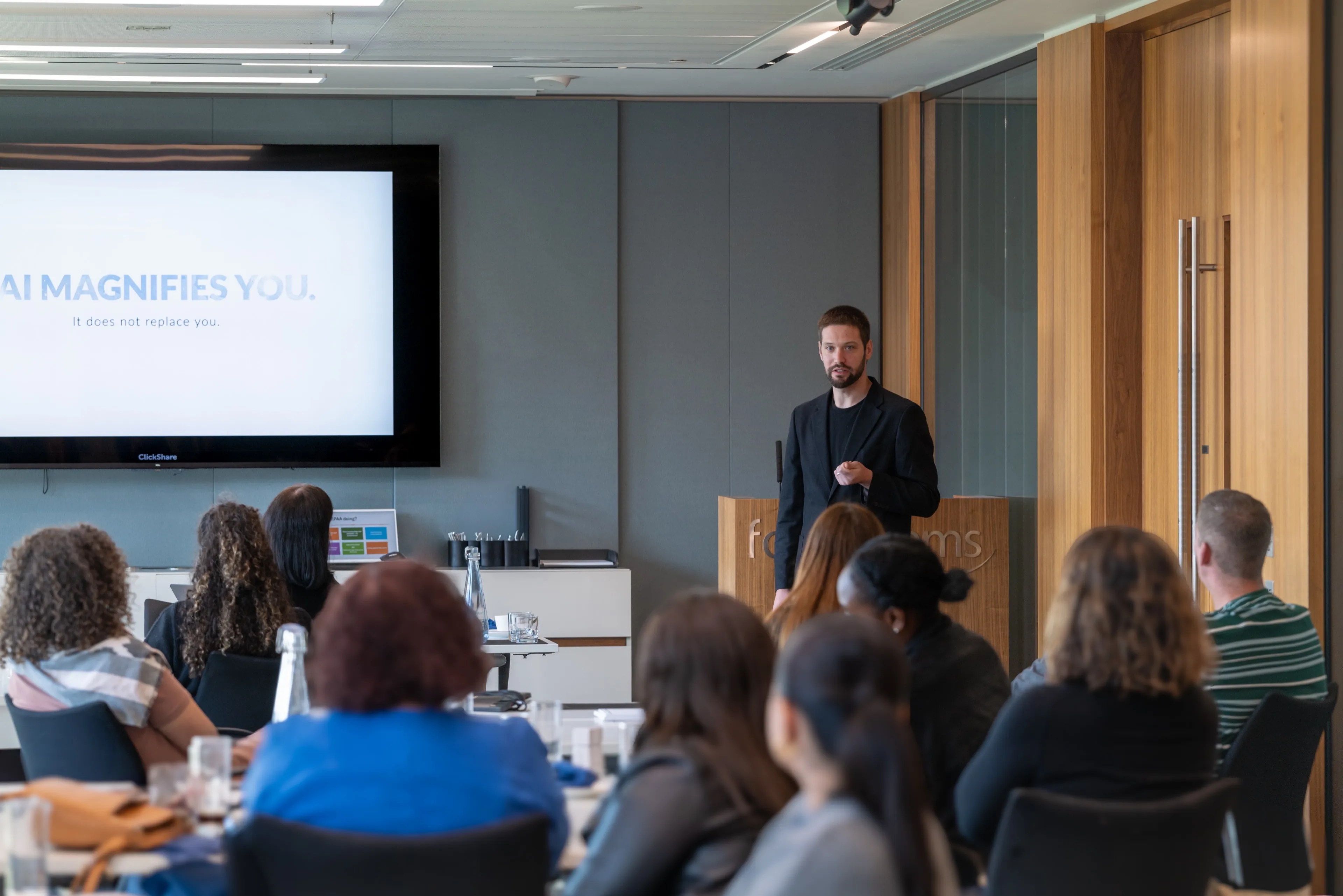 Zach in a black suit giving a presentation to an audience in a modern conference room. A screen behind him reads 'AI MAGNIFIES YOU. It does not replace you.'