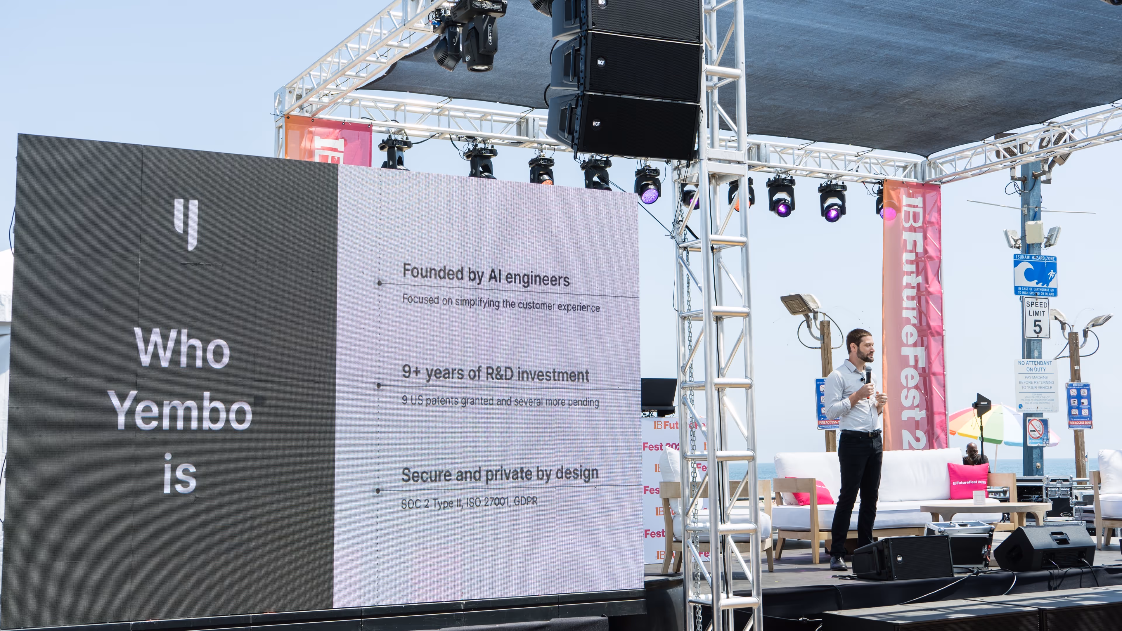 A male speaker presenting on an outdoor stage beside a large digital screen displaying the slide 'Who Yembo is' with key company facts.