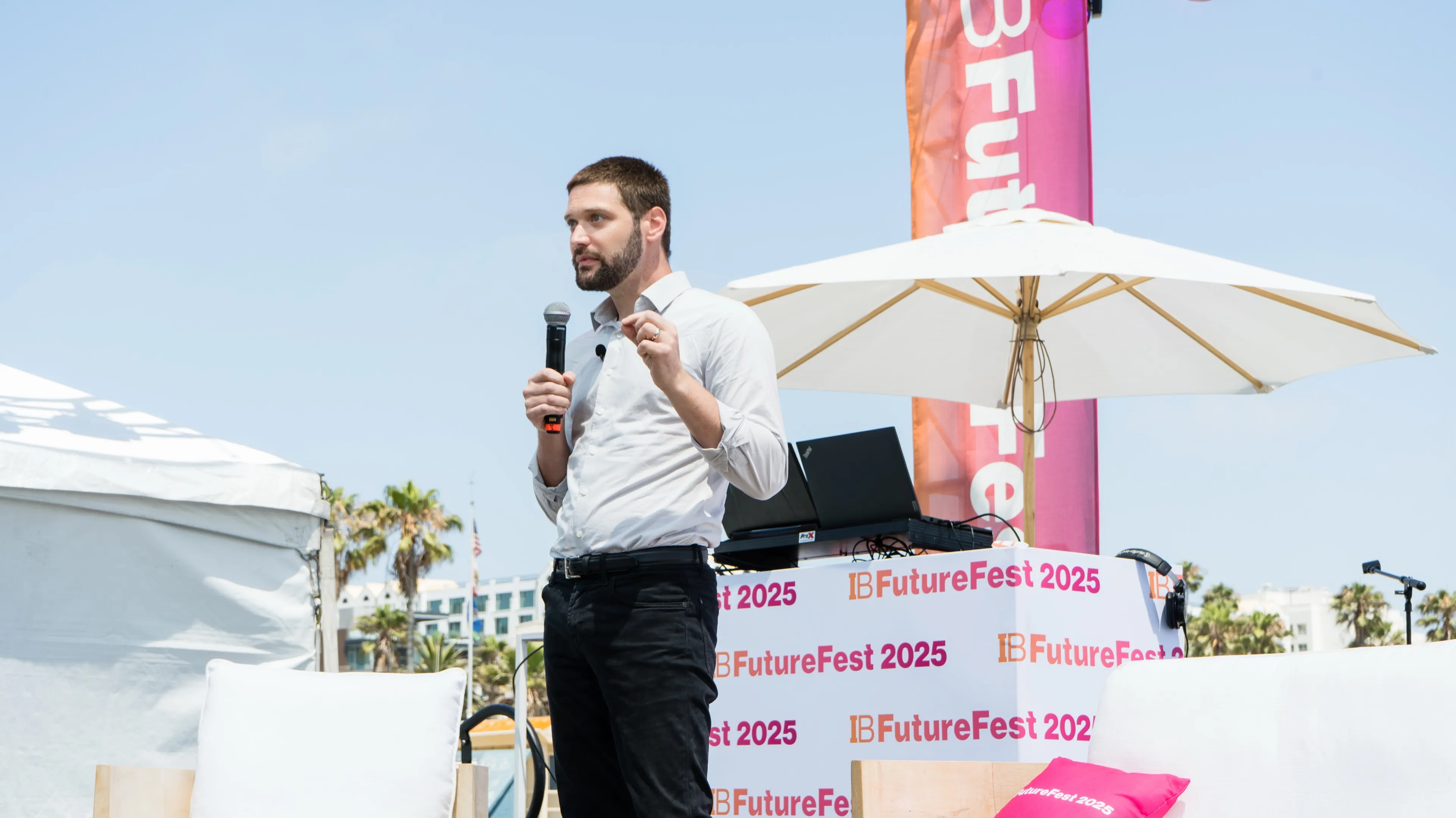 A man with a beard is speaking into a microphone on an outdoor stage with IB FutureFest 2025 branding behind him.