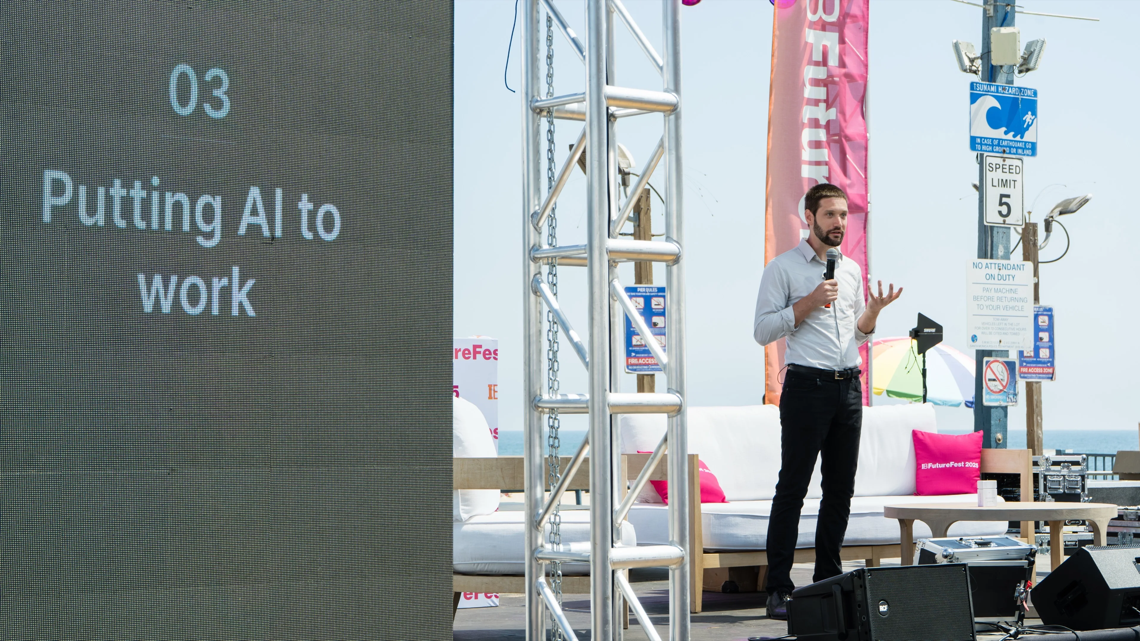 Zach delivering a presentation on an outdoor stage next to a large digital screen displaying the text '03 Putting AI to work'.