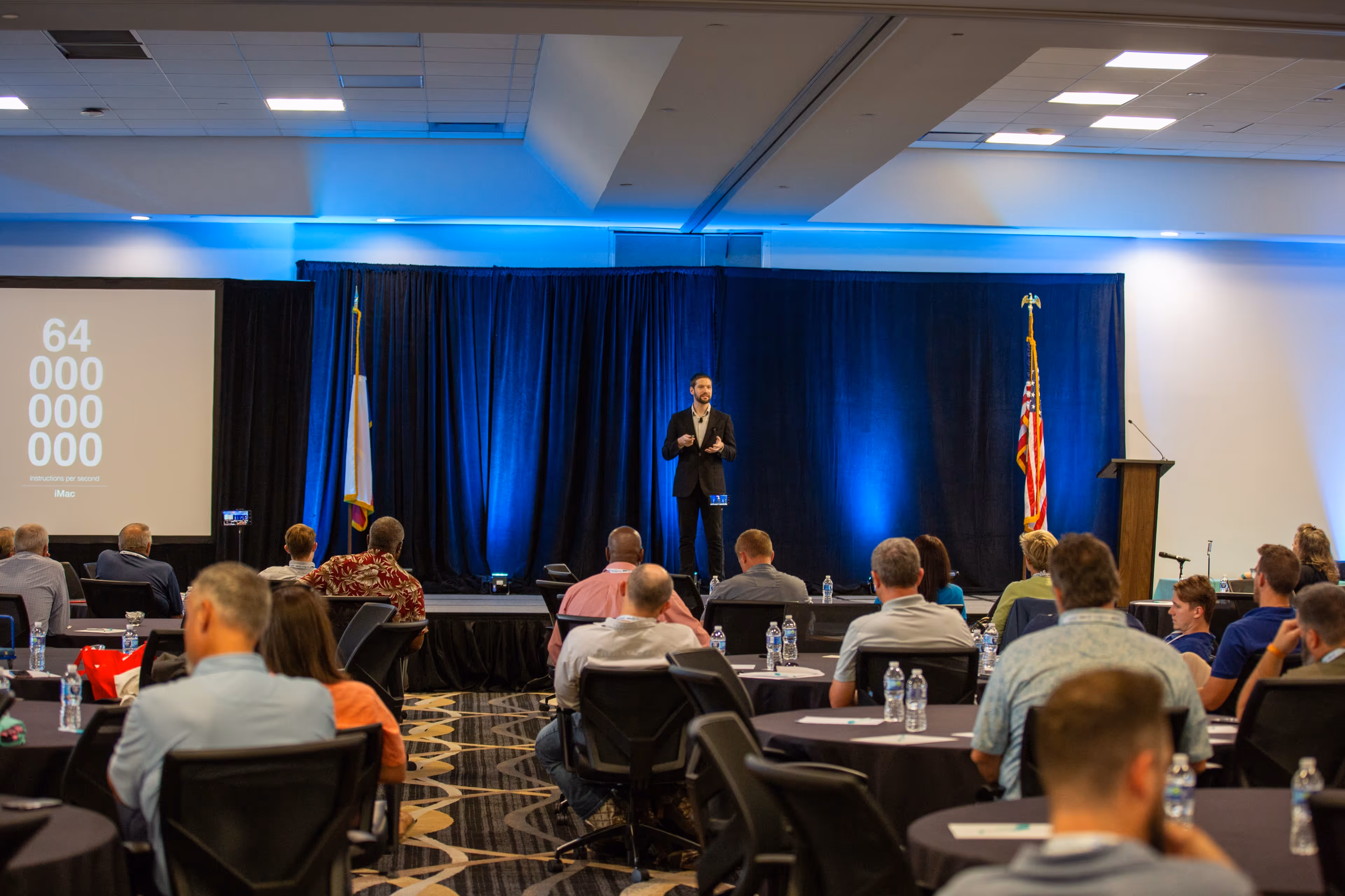 Zach giving a presentation on stage to an attentive audience seated at round tables during a corporate conference event.