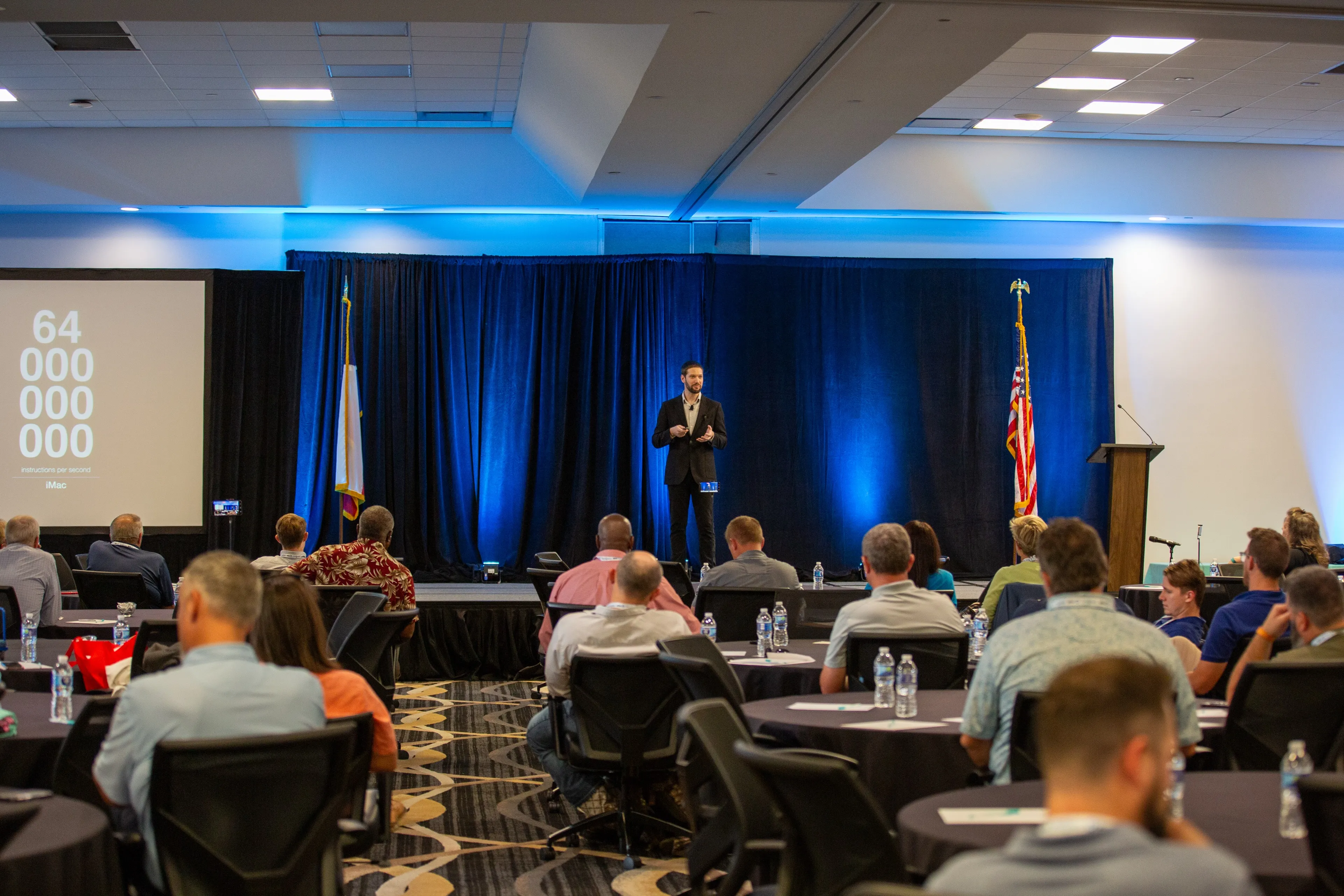 Zach delivering a presentation on a stage to an audience seated at round tables, with a large screen displaying the number 64,000,000,000.