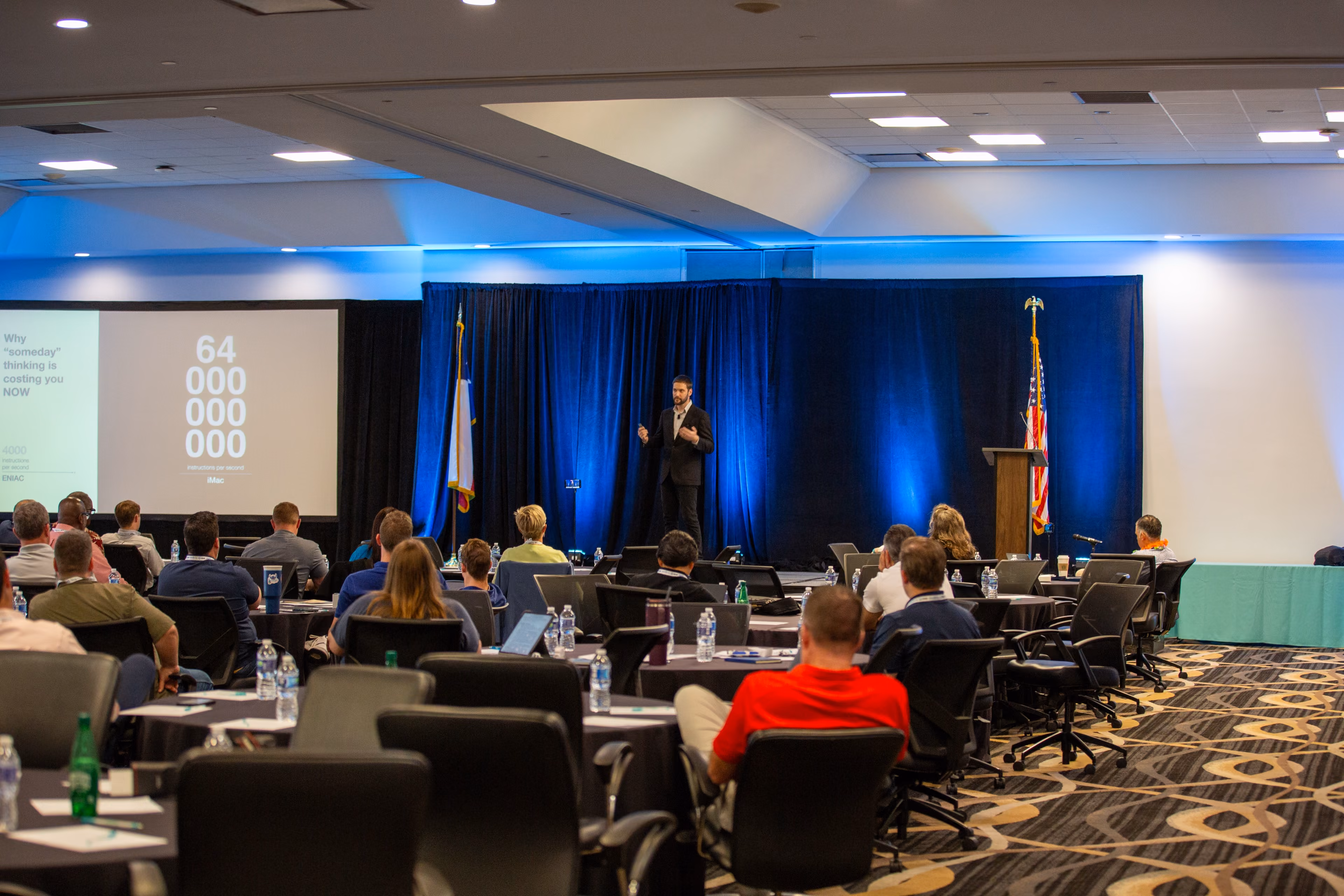 Zach delivering a keynote presentation on a stage with a blue curtain backdrop to an attentive audience seated at round tables.