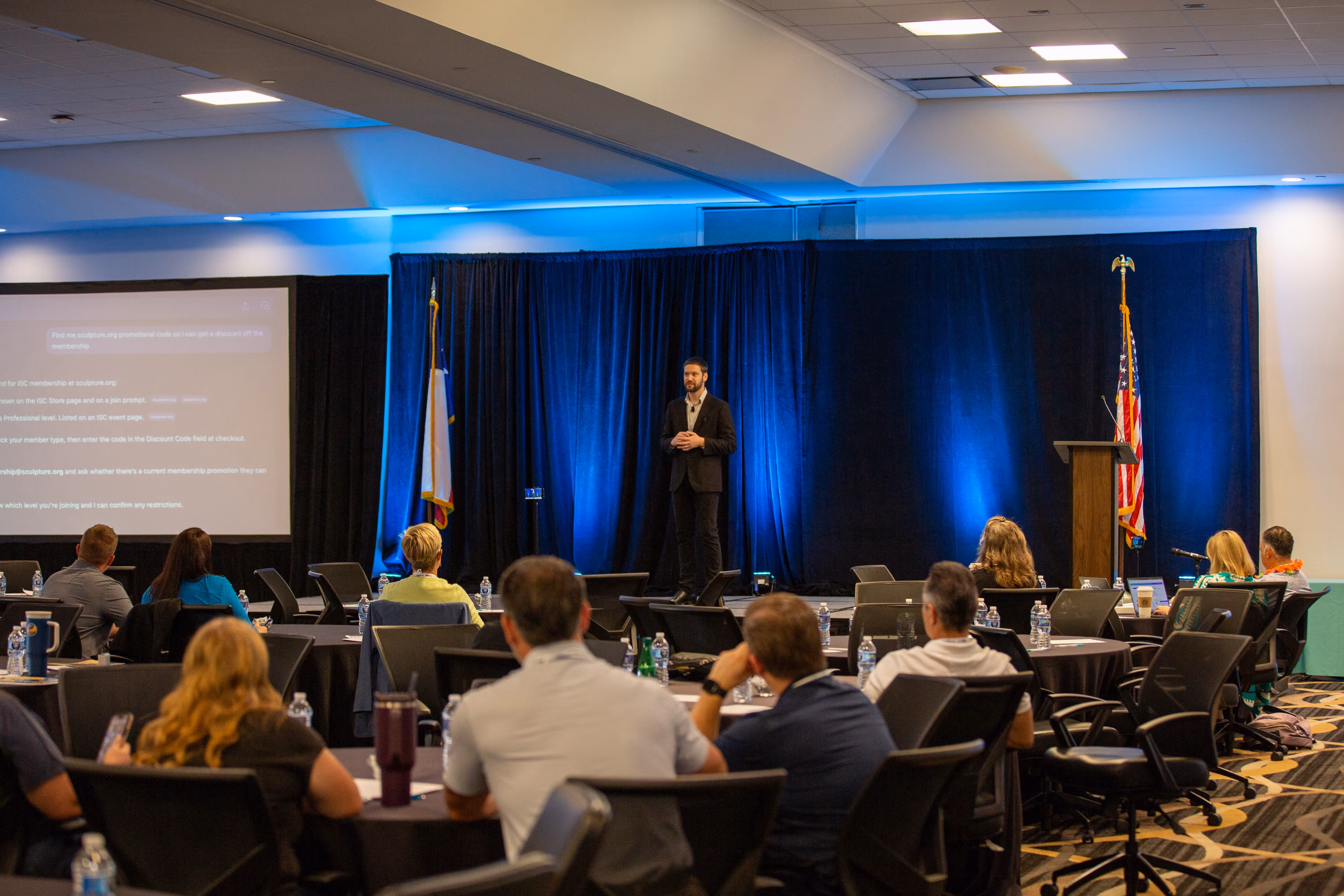 Zach presenting on stage with a blue curtain backdrop to an attentive audience seated at round tables during a corporate conference.