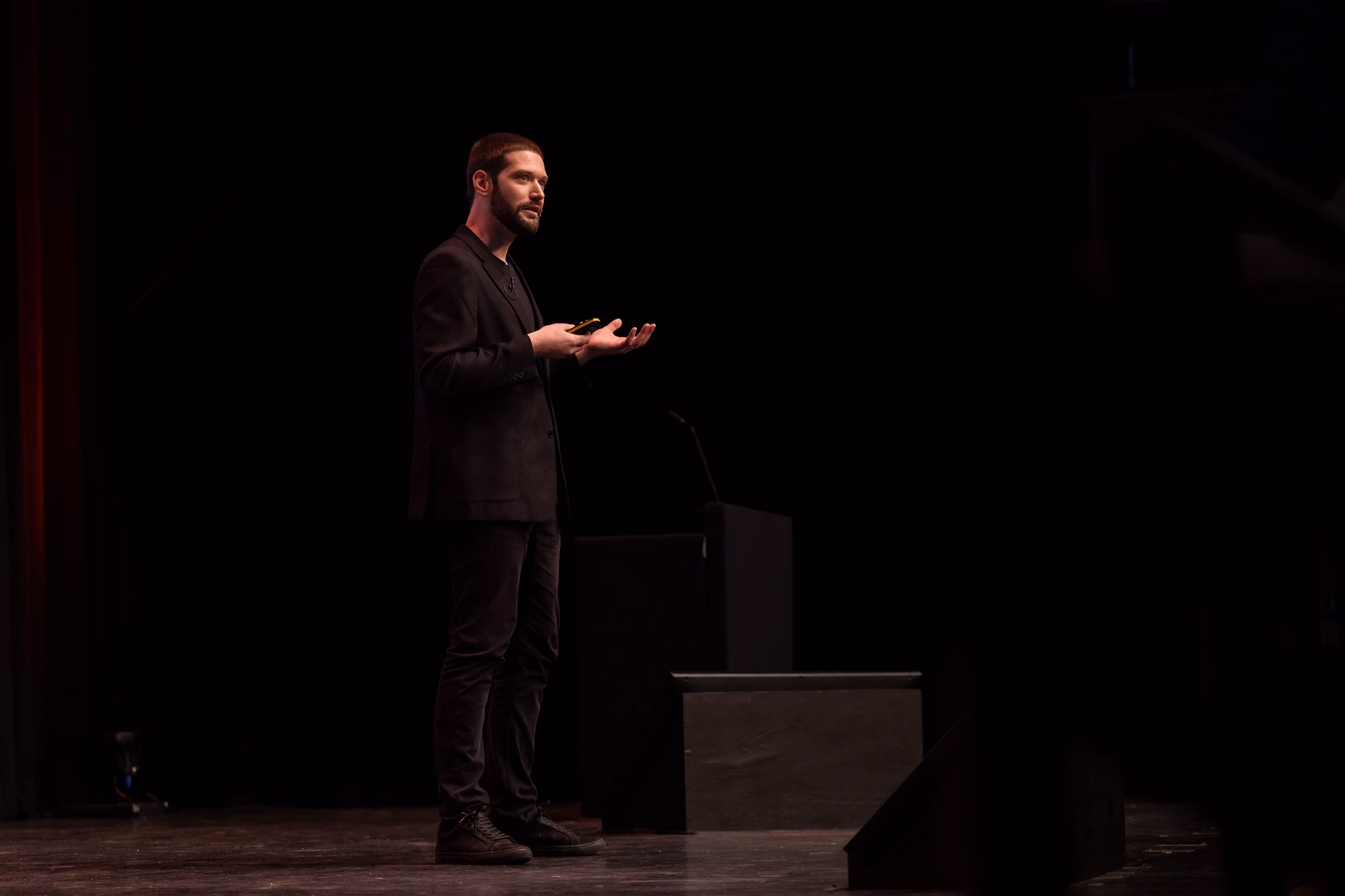 Zach delivering a keynote presentation on a dark stage, gesturing with one hand and holding a clicker.