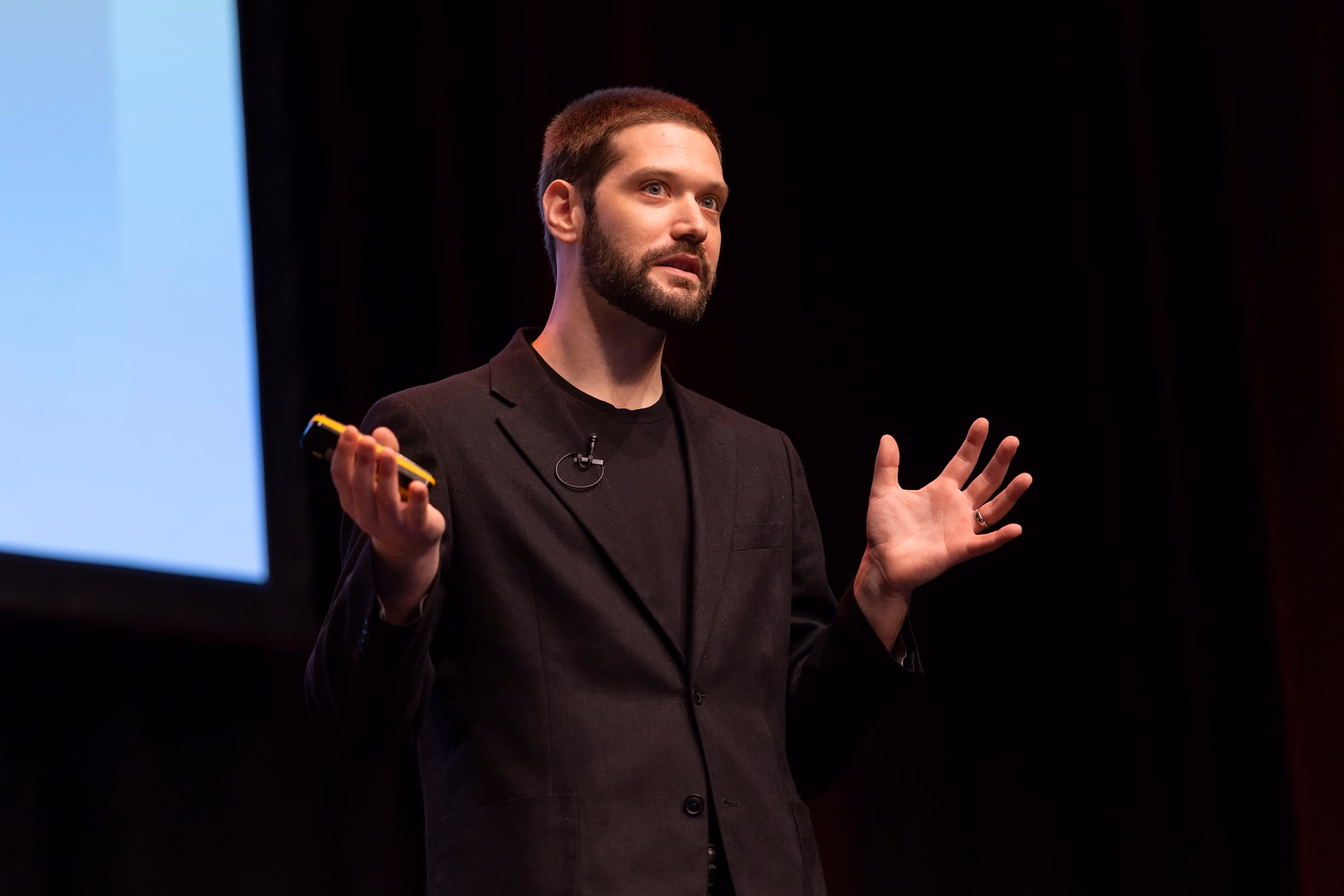 Zach on a stage gesturing with his hands while holding a presentation clicker, wearing a black blazer and lapel microphone with a projection screen partially visible in the background.