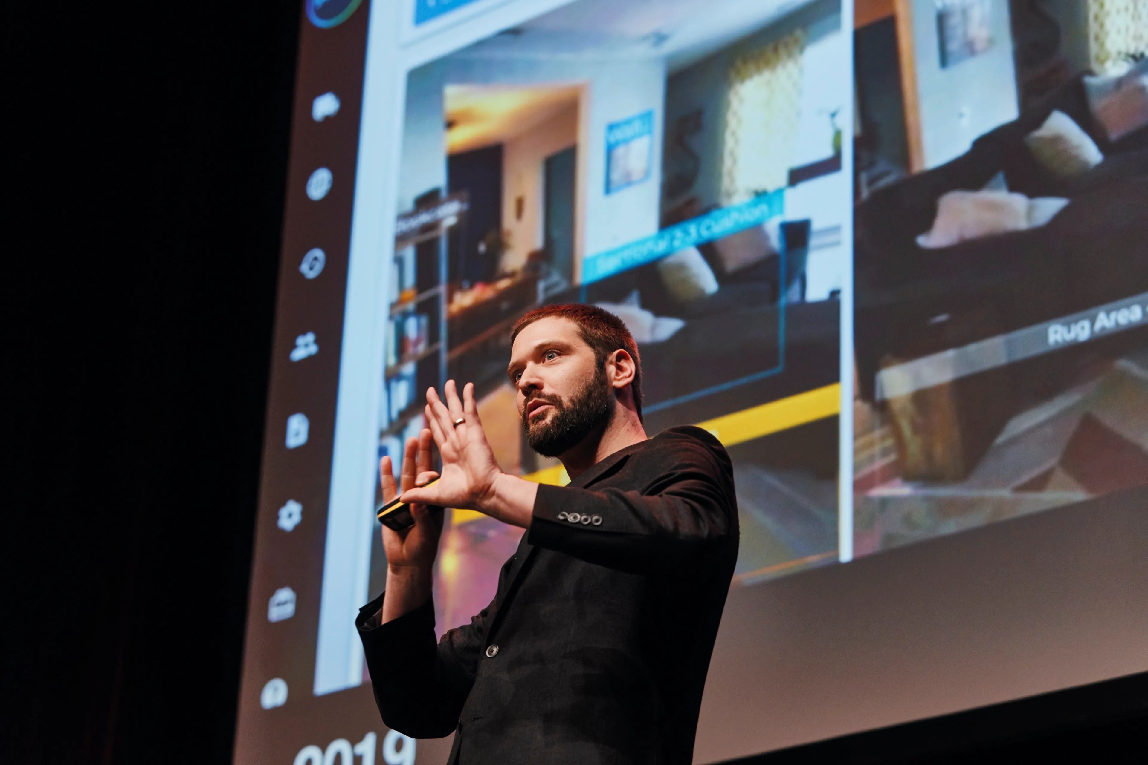 A male speaker on stage actively gesturing with his hands during a tech presentation, with a large screen behind him displaying computer vision or AR technology identifying furniture in a room.