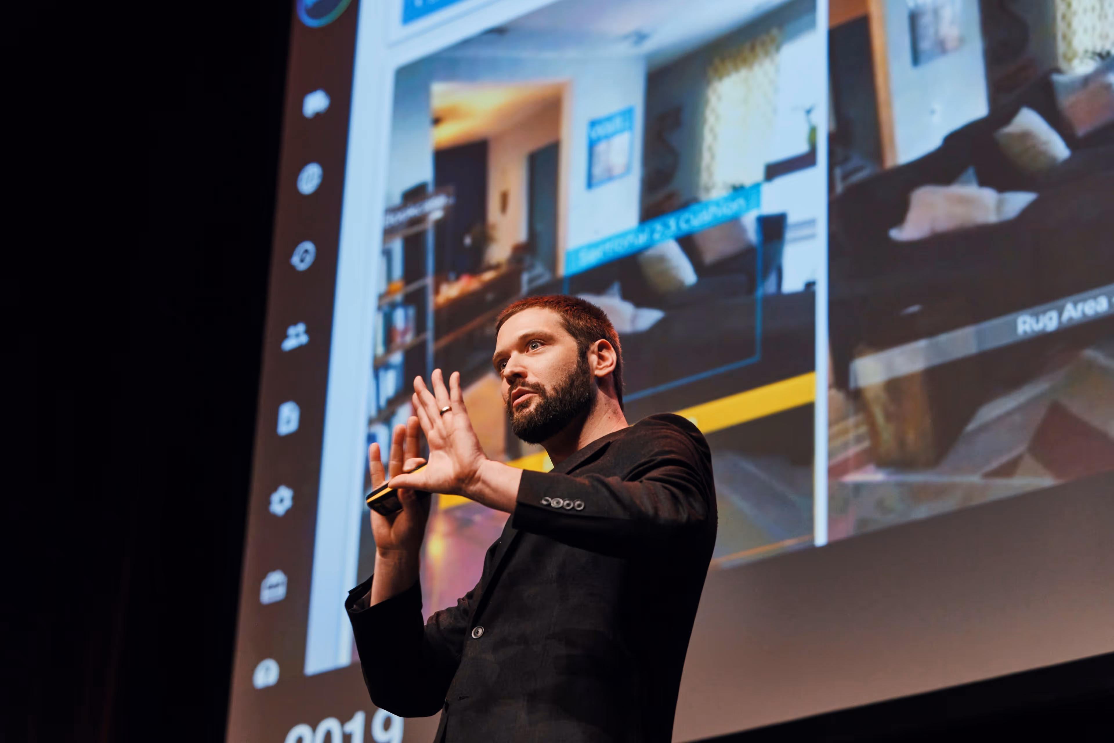 Zach on stage actively gesturing with his hands during a tech presentation, with a large screen behind him displaying computer vision or AR technology identifying furniture in a room.