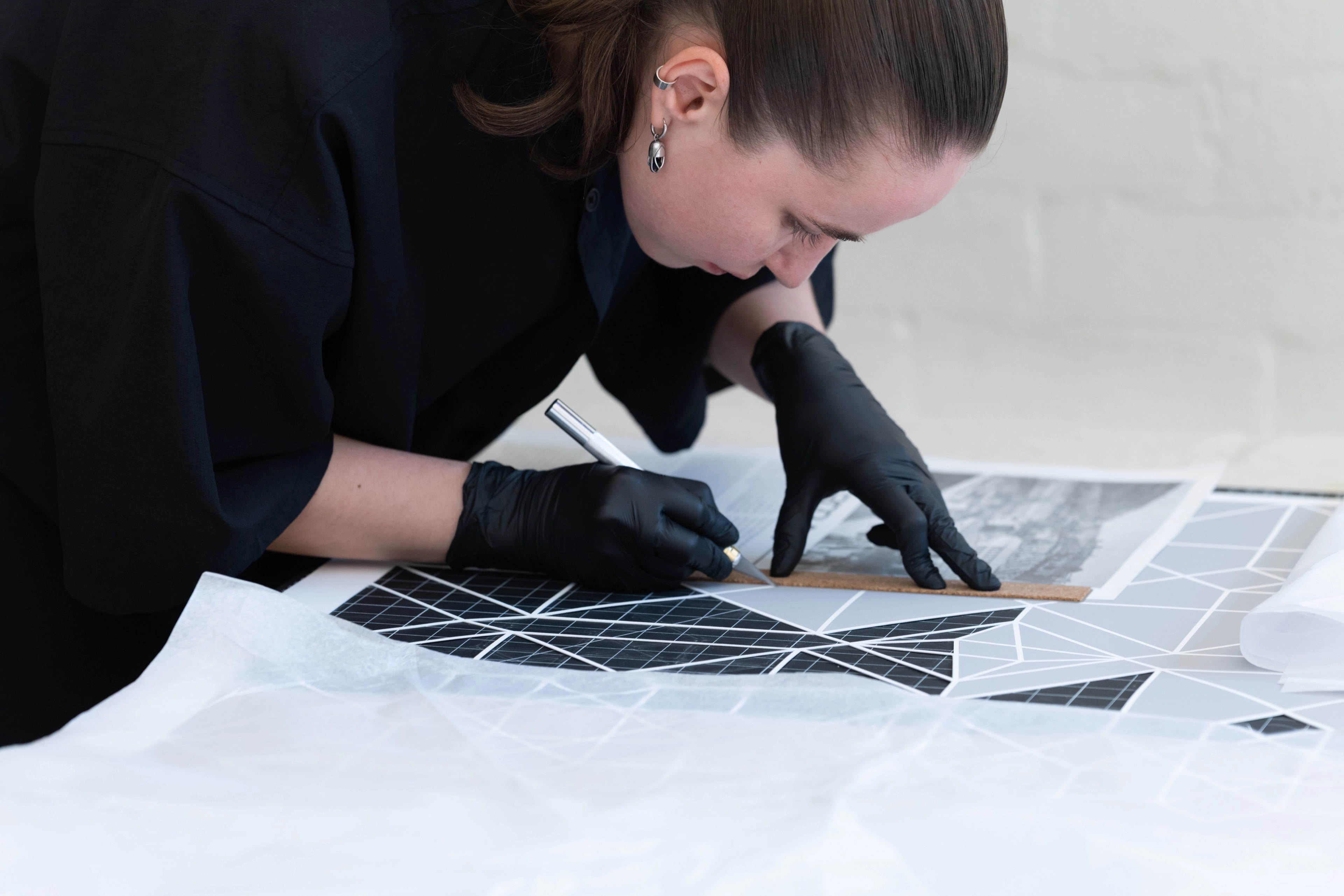 A focused individual wearing black gloves carefully cuts material using an exacto knife and ruler on a geometric cutting mat.