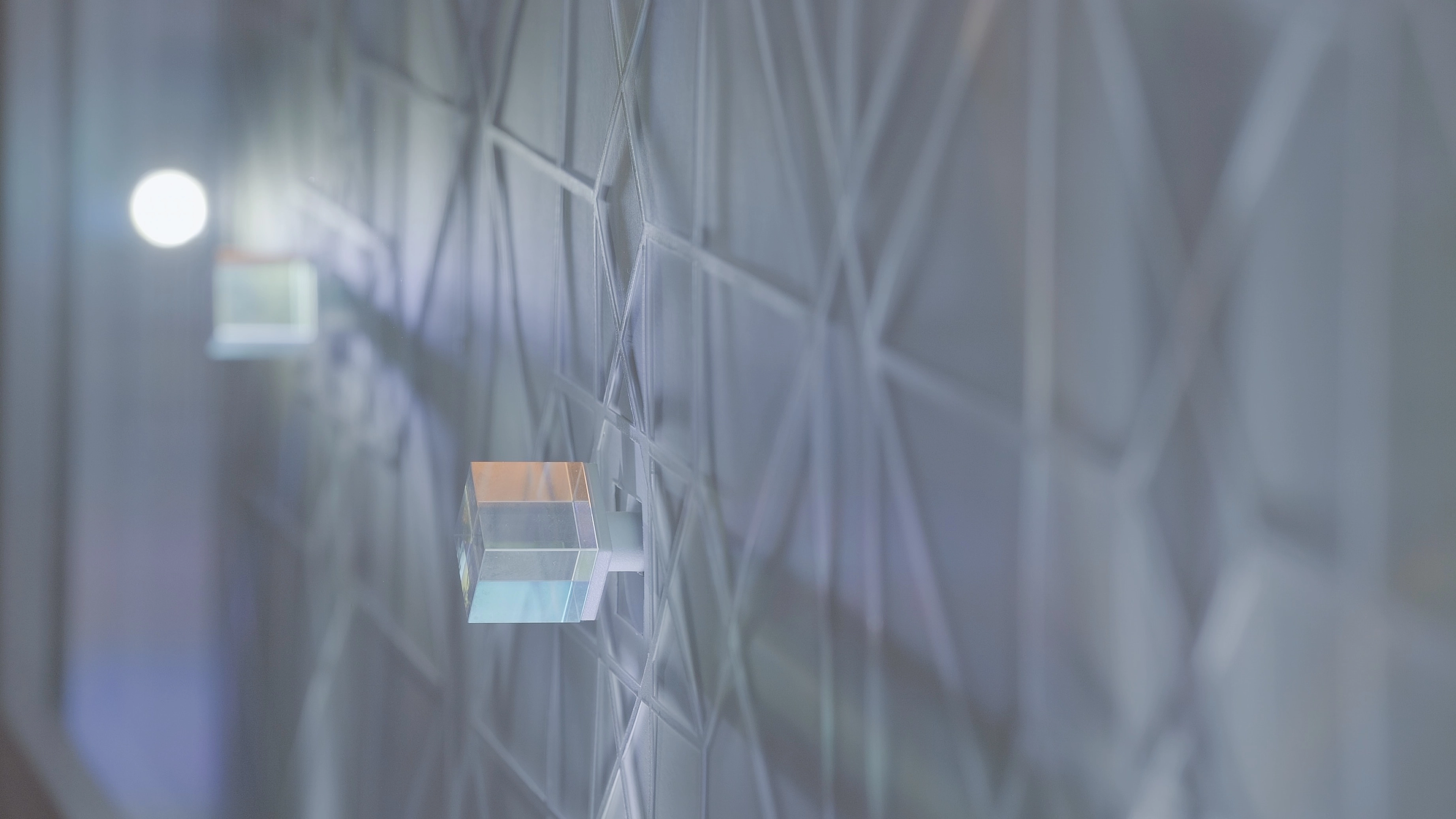 A close-up of a textured white wall featuring an geometric pattern. Two small, translucent cubic light fixtures are mounted on the wall, one in focus in the foreground and another blurred in the background.