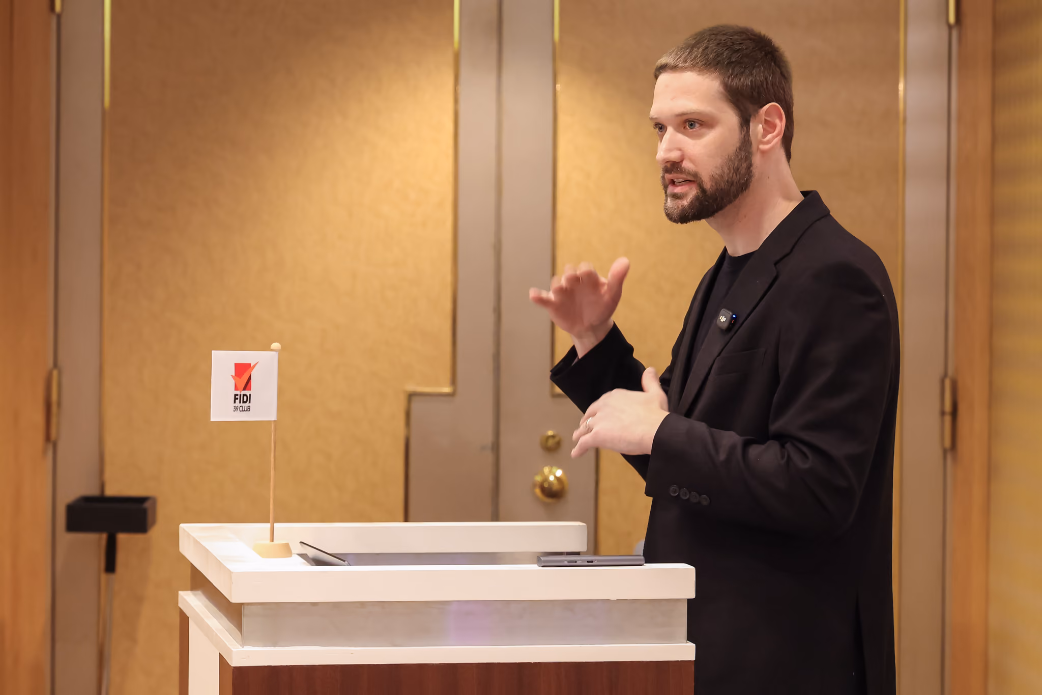 Zach speaking from a white podium topped with a small FIDI 39 Club table flag, gesturing with both hands in front of a gold paneled conference room wall.