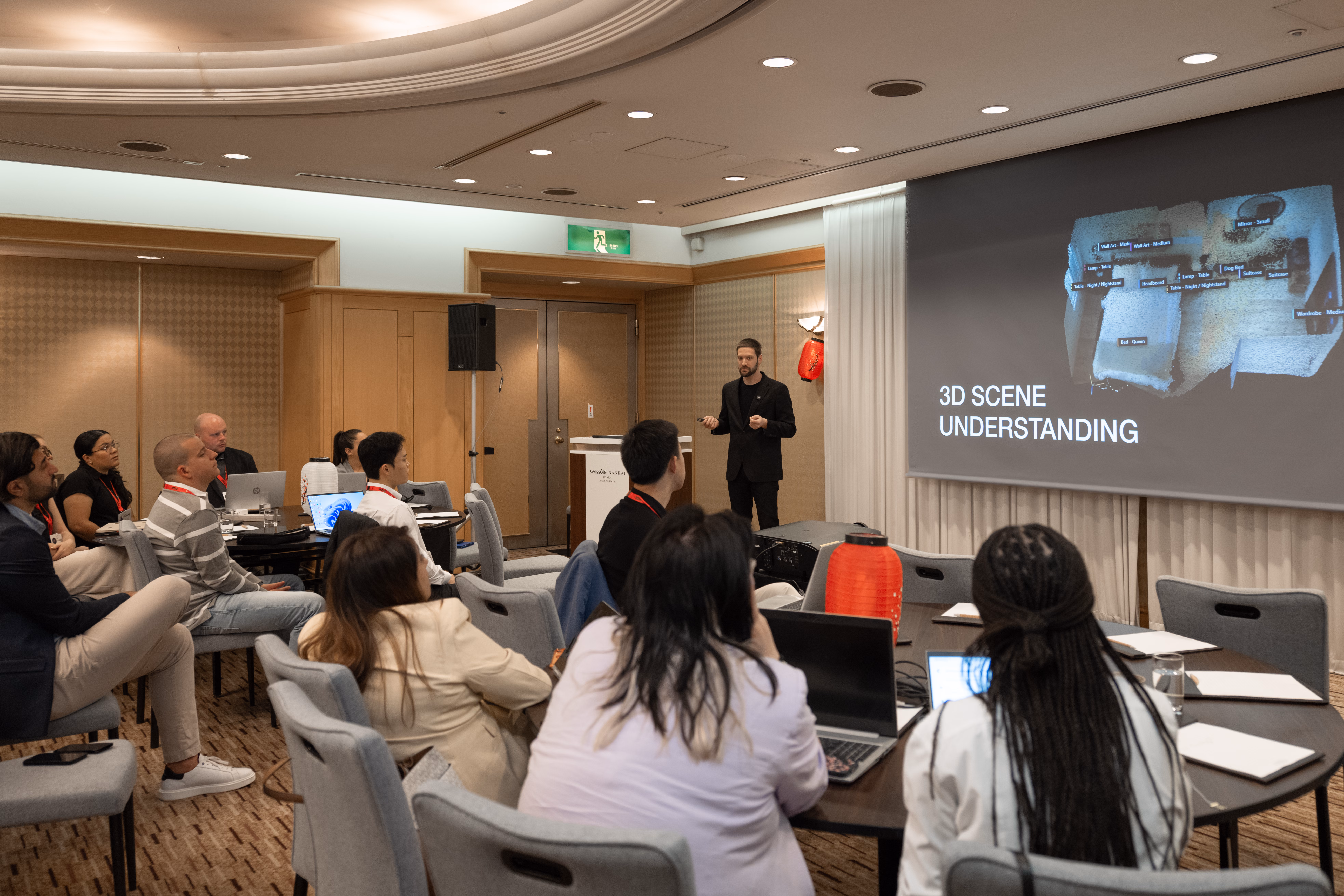 A wide view of a conference room where Zach presents a slide titled 3D Scene Understanding showing a labeled 3D room scan, with attendees seated at tables working on laptops in the foreground.
