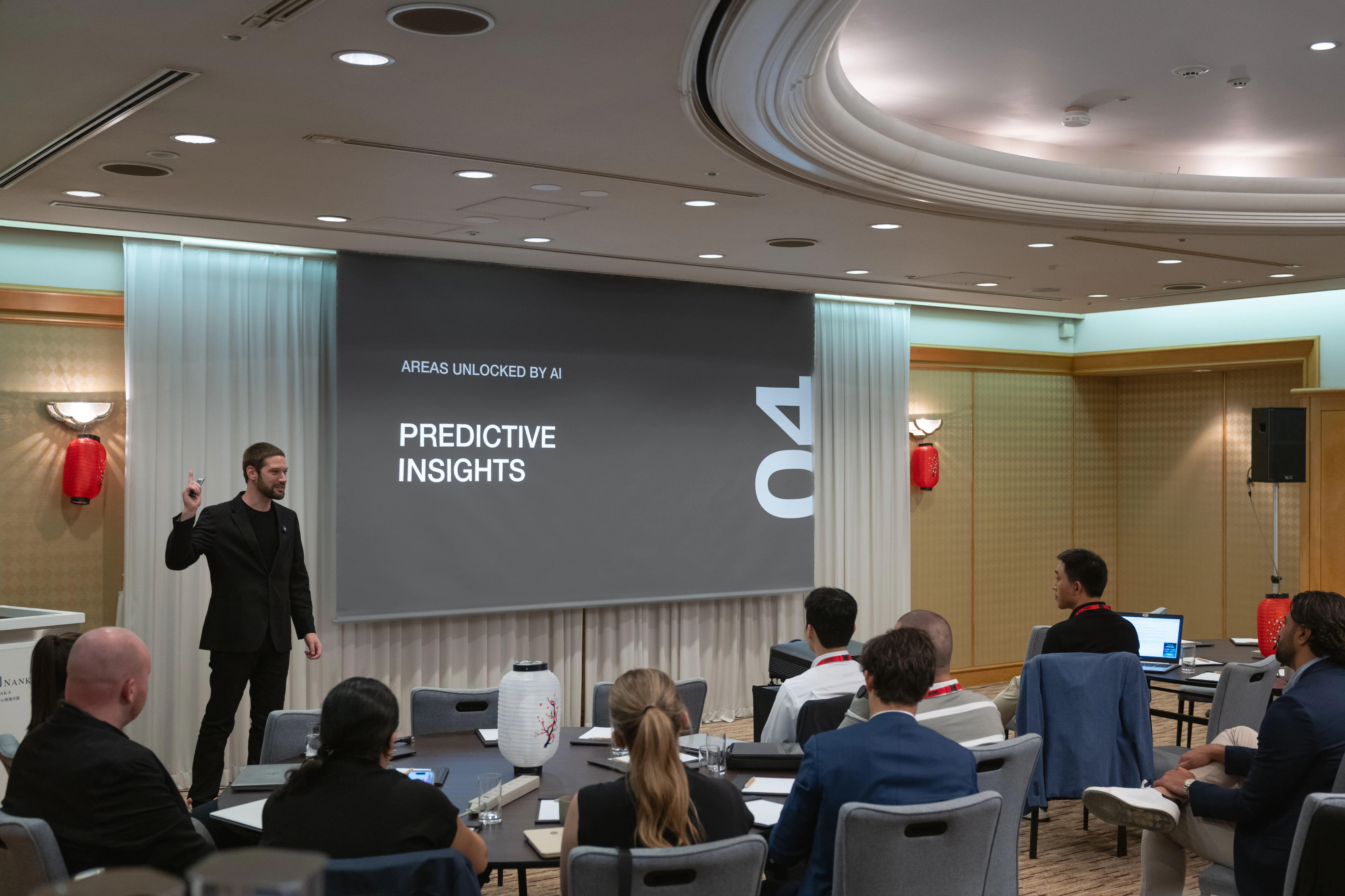 Zach presents beside a large screen displaying a slide titled Predictive Insights under the heading Areas Unlocked by AI, in an ornate conference room decorated with red Japanese lanterns.