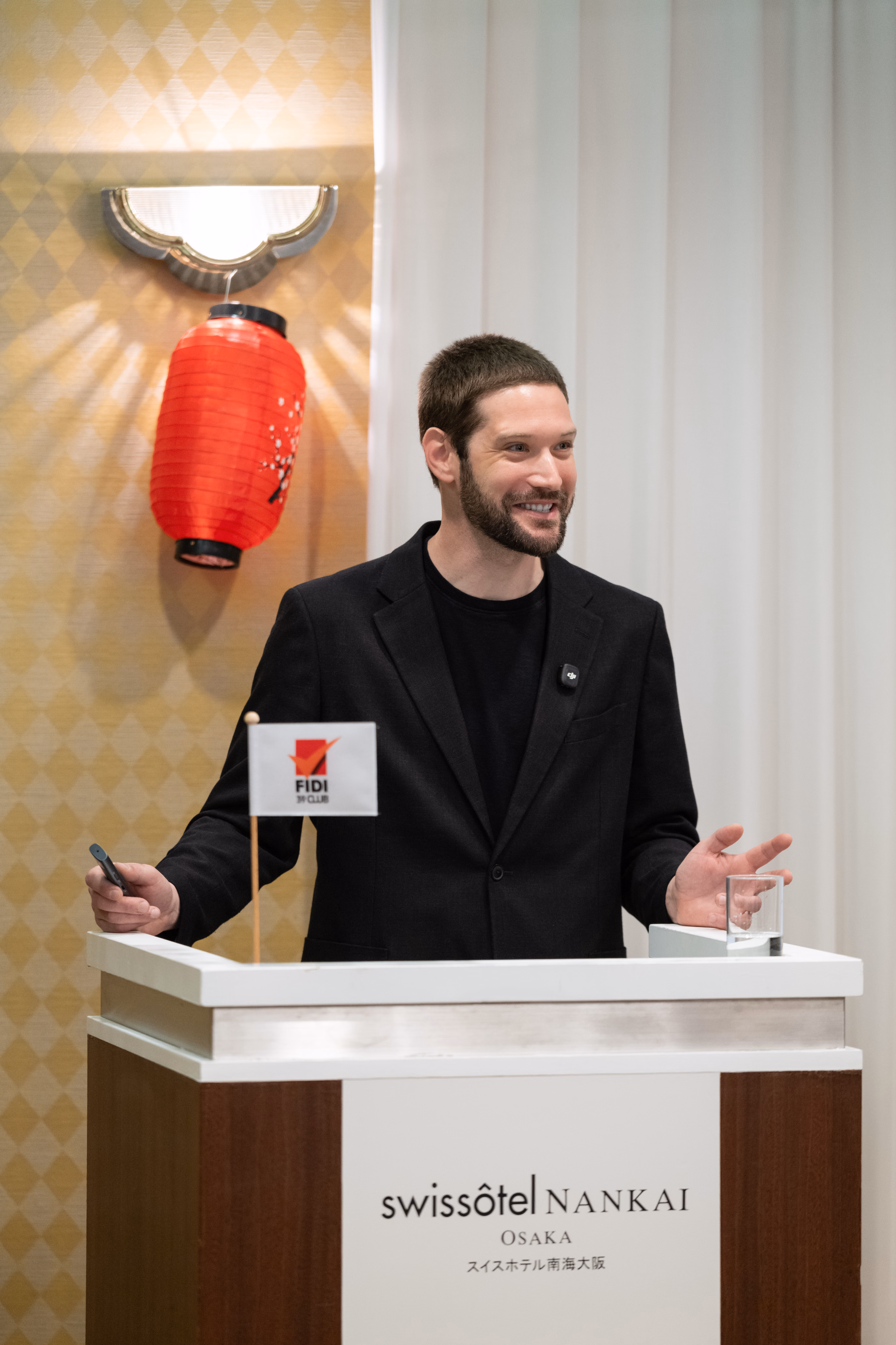 Zach smiles warmly from behind a podium branded Swissotel Nankai Osaka, flanked by a FIDI 39 Club table flag and a red Japanese paper lantern beneath a wall sconce on a gold patterned wall.