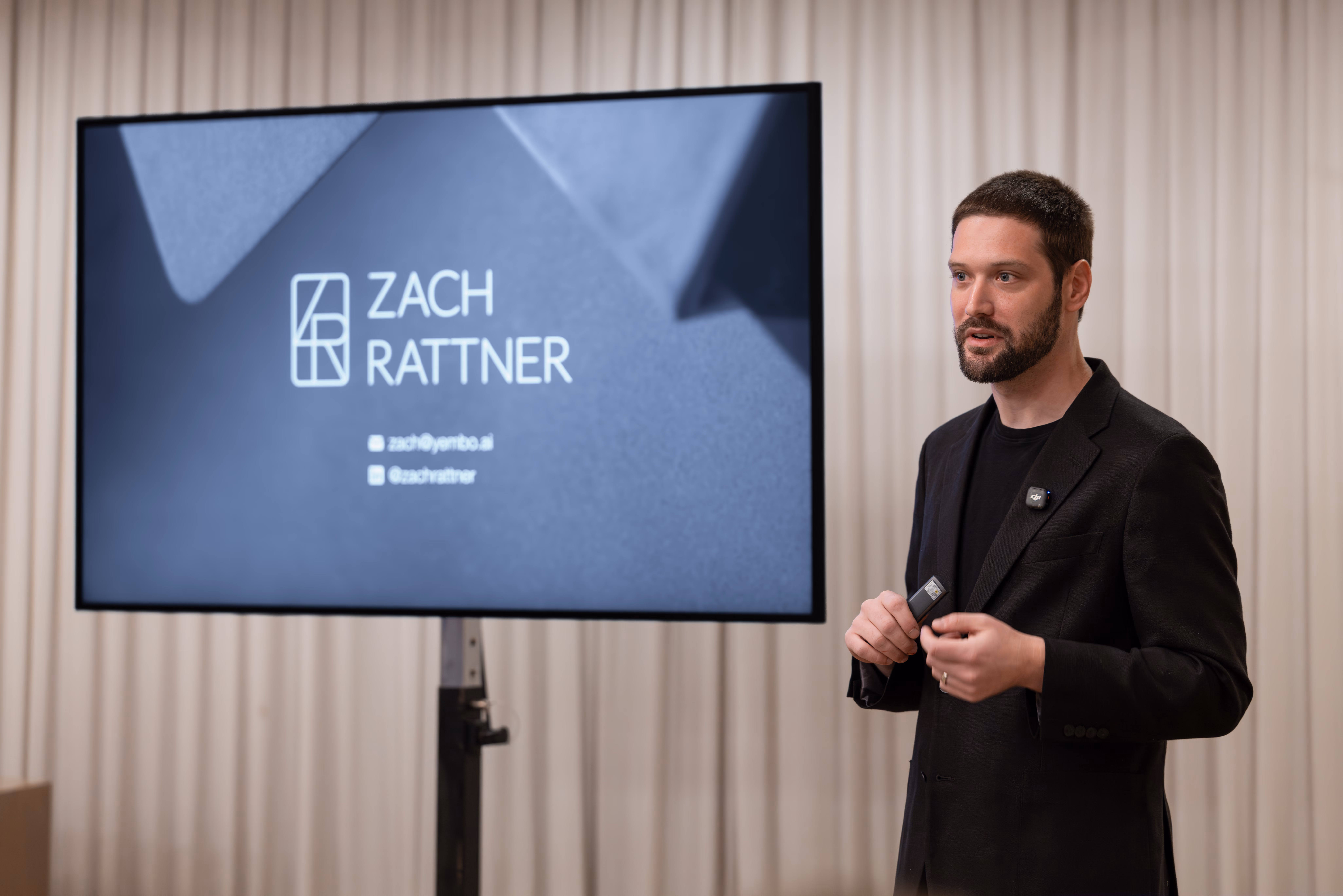 Zach presents beside a display showing his personal title slide with the Zach Rattner logo and contact details, holding a presenter remote in both hands against a cream curtain backdrop.